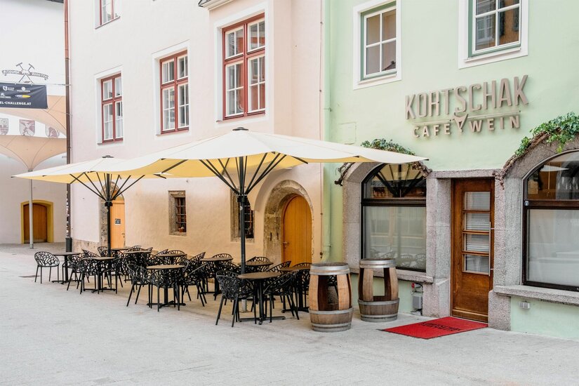 Außenansicht des Café Kortschak mit Tischen, Stühlen und Sonnenschirmen in einer gepflasterten Gasse.