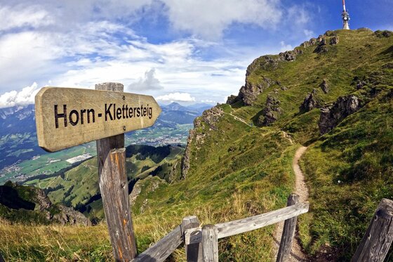 Wegweiser Horn-Klettersteig vor alpinem Wanderweg und Sendemast auf einem Berggipfel.