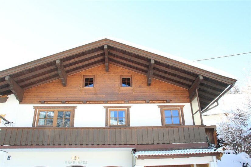 Vorderansicht eines zweistöckigen Alpenhauses mit Holzbalkon und schneebedecktem Satteldach.