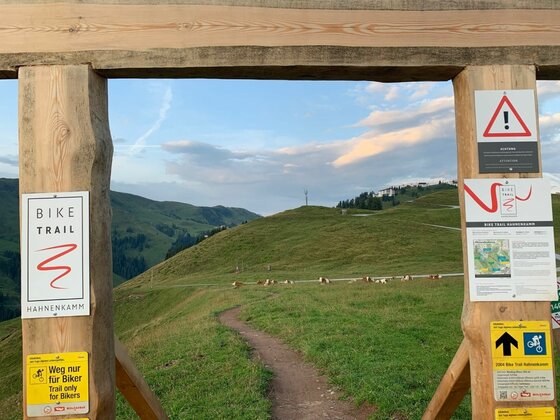 Start Bike Trail Hahnenkamm Holztor mit Hinweisschildern am Start des Bike Trail Hahnenkamm vor alpiner Berglandschaft.
