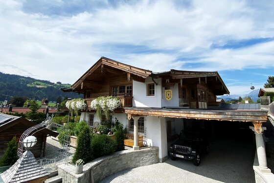 Alpines Wohnhaus aus Holz und Stein mit Blumenbalkonen und Carport vor Bergkulisse.