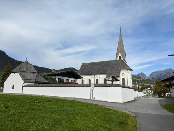 Pfarrkirche in Reith Weiße Kirche mit spitzem Turm und umlaufender Mauer vor bewaldeten Bergen.