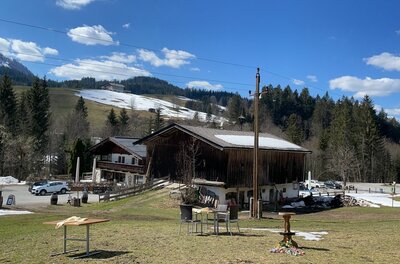 Alpines Holzgebäude mit Parkplatz und Außenbestuhlung vor bewaldeten Bergen mit Schneeresten.