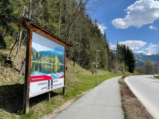Große Informationstafel an einem asphaltierten Rad- und Gehweg neben einer Landstraße vor bewaldetem Hang.