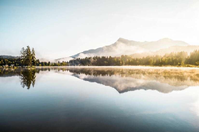 Ein ruhiger See mit Spiegelung der Berge und Nadelbäume bei leichtem Morgennebel.