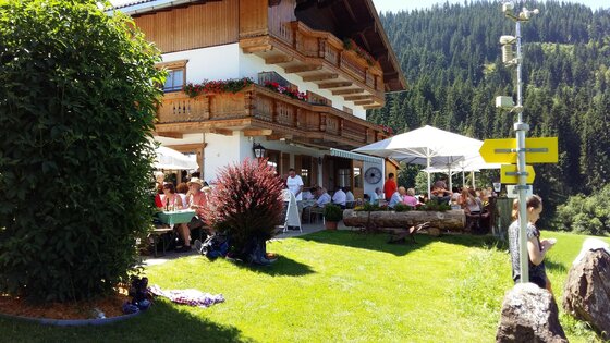 Ein gemütliches Restaurant mit Sonnenschirmen und Gästen im Freien, umgeben von grünen Wiesen und Bergen. Die Fassade des Hauses ist im traditionellen Stil mit Blumenkästen dekoriert.