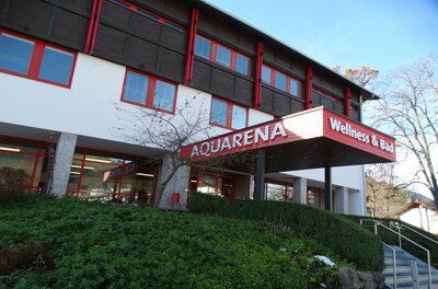 Das Gebäude hat ein rotes Schild mit der Aufschrift "AQUARENA Wellness & Bad". Es ist von einer grünen Landschaft umgeben.