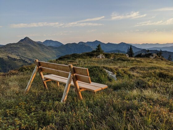 Rastbank am Gebra Holzbank auf einer Bergwiese mit Blick auf ein Gebirgspanorama bei Sonnenuntergang.