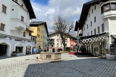 Gepflasterter Dorfplatz mit einem Steinbrunnen im Zentrum, umgeben von mehrstöckigen Häusern mit hellen Fassaden.