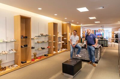 Zwei Männer in einem hellen Schuhgeschäft mit Holzregalen und verschiedenen Schuhmodellen.