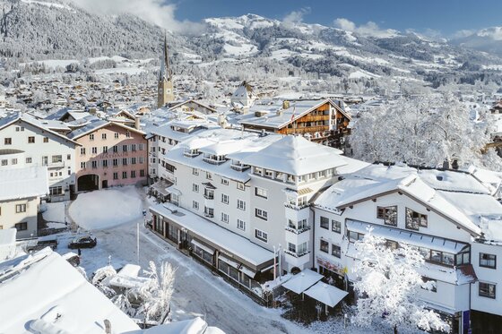 Eine winterliche Stadtansicht mit schneebedeckten Dächern und bergiger Kulisse. Die Architektur der Häuser ist vielfältig und die Landschaft strahlt eine ruhige Atmosphäre aus.