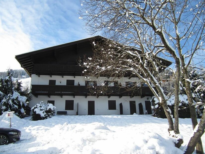 Mehrstöckiges weißes Gebäude mit Holzbalkonen in einer verschneiten Winterlandschaft unter blauem Himmel.
