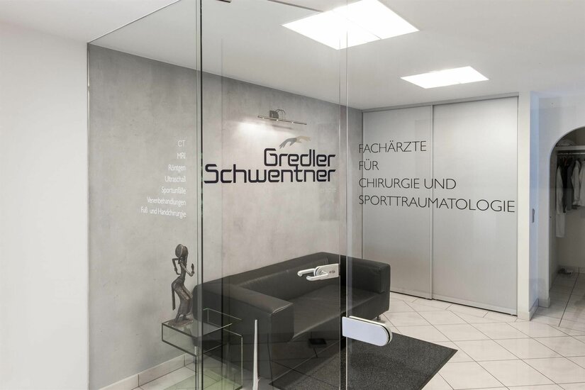 Praxis Dr. Gredler und Schwentner Eingangsbereich einer Arztpraxis mit Glastür, schwarzem Sofa und Beschriftung an der Wand.