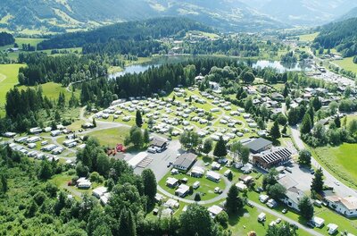 Luftaufnahme eines Campingplatzes mit Wohnwagen und Zelten an einem See zwischen bewaldeten Hügeln.