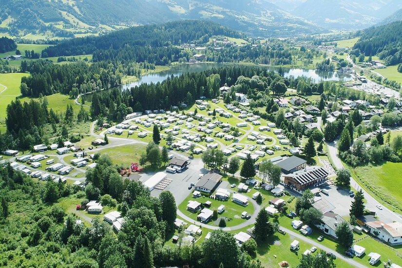 Luftaufnahme eines Campingplatzes mit Wohnwagen und Zelten an einem See zwischen bewaldeten Hügeln.