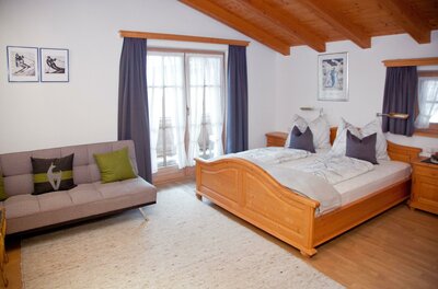 Schlafzimmer mit Doppelbett aus Holz, grauem Sofa und Balkontür unter einer Holzdecke.