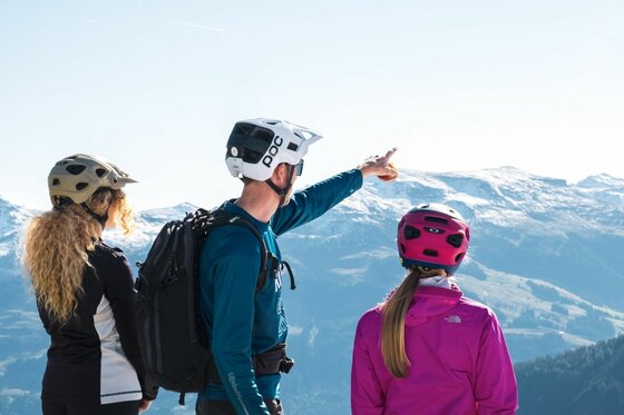 Mountainbiker mit Bergpanorama Drei Mountainbiker mit Helmen blicken von einem Gipfel auf schneebedeckte Berge.
