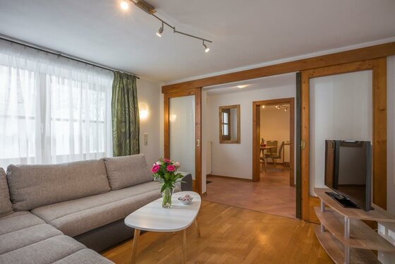 Wohnbereich im Appartement Wetti Wohnzimmer mit grauem Ecksofa, hellem Couchtisch und Fernseher vor einer hölzernen Schiebetür.