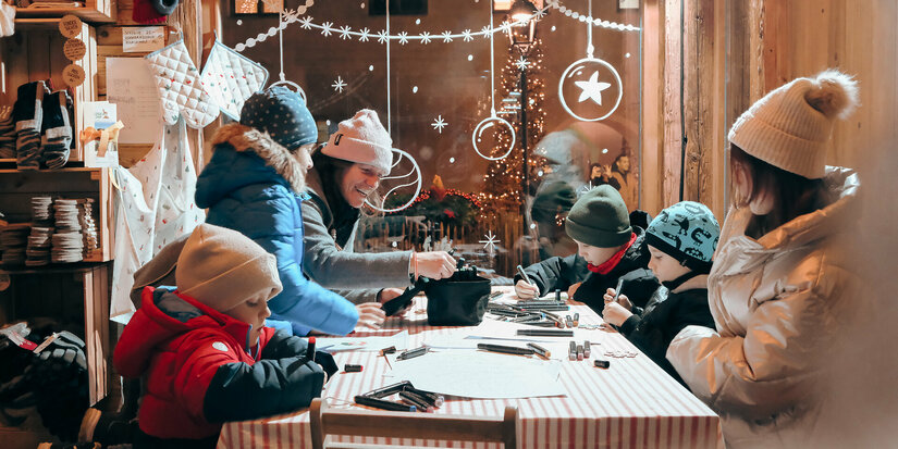 Eine fröhliche Gruppe von Kindern und Erwachsenen bastelt während der Adventszeit in einer gemütlichen Holzhütte in Kitzbühel.