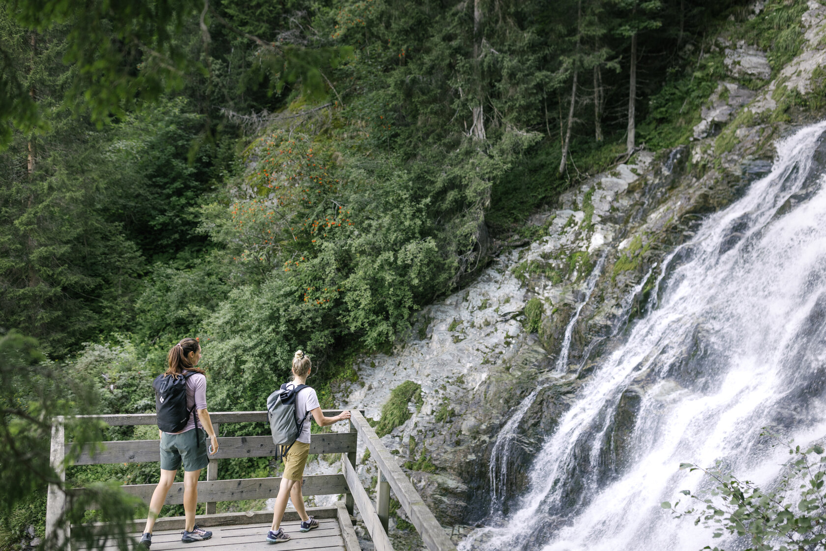 Two women are standing on a viewing platform in front of the Sintersbach Waterfall in Jochberg
