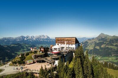 Berghütte mit großer Terrasse auf einem Gipfel vor alpinem Bergpanorama bei blauem Himmel.