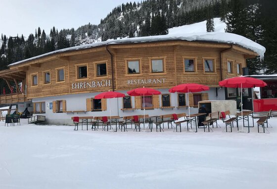 Ehrenbachgraben Winter Holzhütte mit Restaurant-Schriftzug, roten Sonnenschirmen und Bierbänken im Schnee vor bewaldetem Hang.