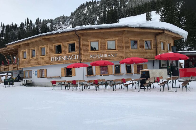 Ehrenbachgraben Winter Holzhütte mit Restaurant-Schriftzug, roten Sonnenschirmen und Bierbänken im Schnee vor bewaldetem Hang.