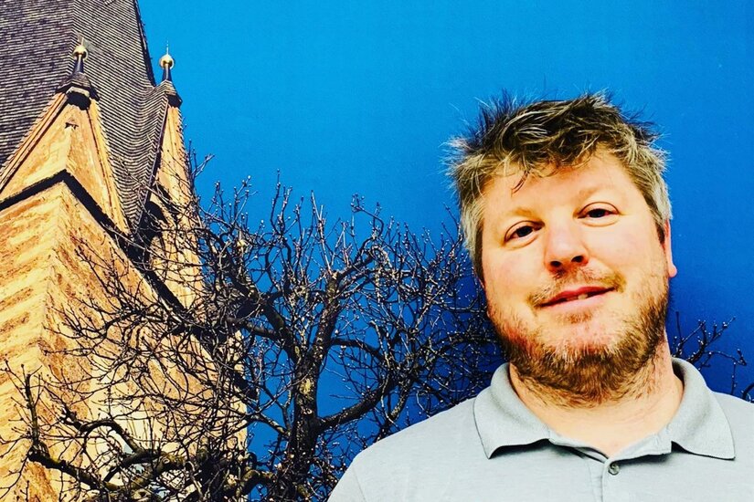 Ein Mann mit Bart vor einem Kirchturm und einem kahlen Baum unter blauem Himmel.
