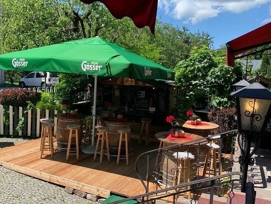 Fritz' LICHTBLICK Ausschank Außenbereich einer Bar mit Holzterrasse, Weinfässern als Stehtische und grünem Sonnenschirm.
