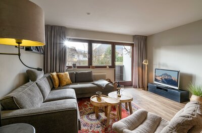 Wohnzimmer Appartement Hafkamp Modernes Wohnzimmer mit grauem Ecksofa, Holztischen, Orientteppich und Fernseher vor einem Fenster mit Bergblick.