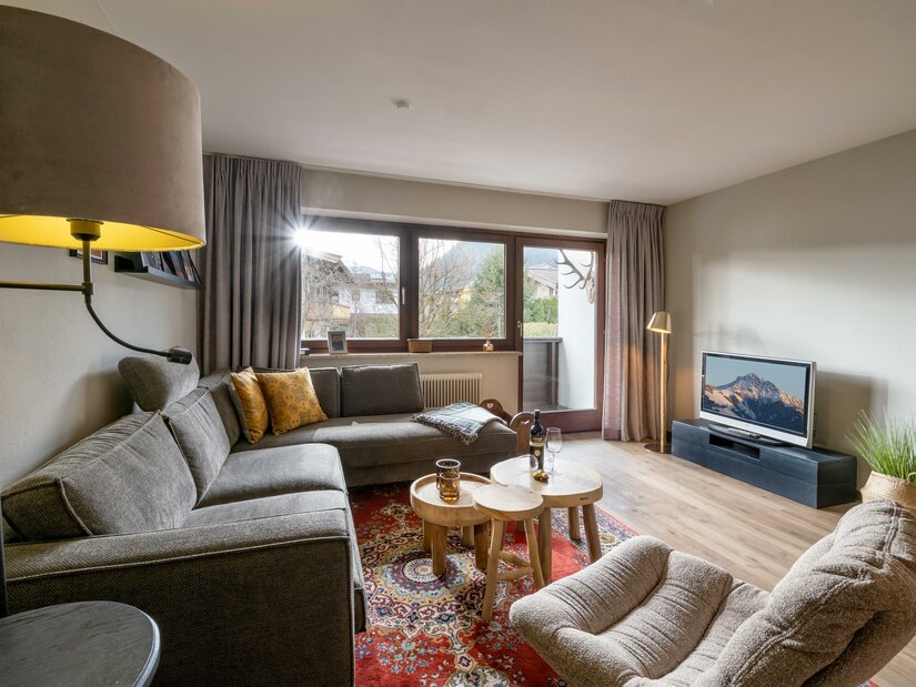 Wohnzimmer Appartement Hafkamp Modernes Wohnzimmer mit grauem Ecksofa, Holztischen, Orientteppich und Fernseher vor einem Fenster mit Bergblick.