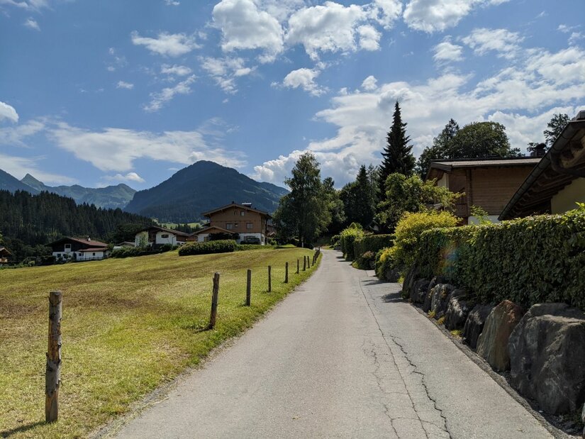 Wanderweg bei Pürsting Asphaltierter Weg führt an einer Wiese und Häusern vorbei auf bewaldete Berge unter bewölktem Himmel zu.