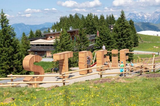 Spielplatz an der Streif Große Holzbuchstaben des Wortes STREIF als Spielplatz vor einer Almhütte und Nadelbäumen.