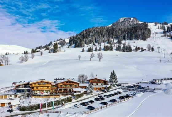 Rosi´s Sonnbergstuben im Winter Verschneite Holzhäuser mit Parkplatz vor bewaldetem Berghang unter blauem Himmel.