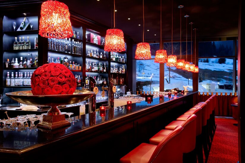 Moderne Hotelbar mit roten Hängelampen, Barhockern und Blick auf eine verschneite Winterlandschaft.