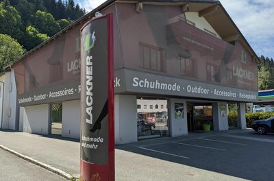 Lackner Schuhe Fachgeschäft Außenansicht des Schuhgeschäfts Lackner mit Werbestele und Parkplätzen vor dem Gebäude.