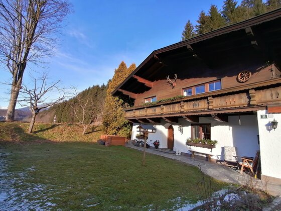Alpines Wohnhaus mit dunkler Holzfassade, Balkon und weißem Erdgeschoss vor Waldhintergrund.