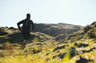 Ein Trailrunner läuft von hinten gesehen über eine hügelige, grüne Almwiese in Richtung eines Bergrückens.