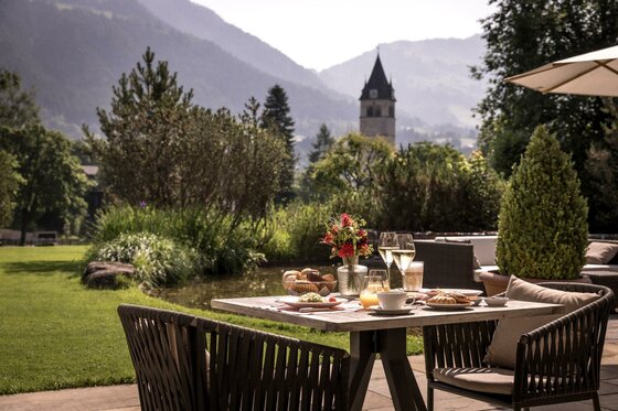 Ein schöner Garten mit einem Tisch, auf dem Frühstück und Getränke serviert werden. Im Hintergrund sind Berge und ein Kirchturm sichtbar.