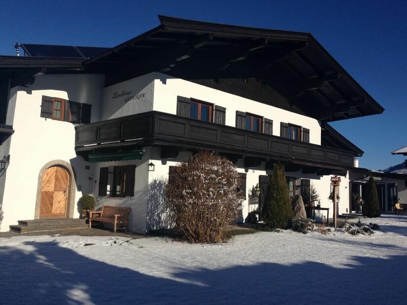 Zweistöckiges weißes Landhaus mit dunklem Holzbalkon und Schneefläche im Vordergrund bei blauem Himmel.
