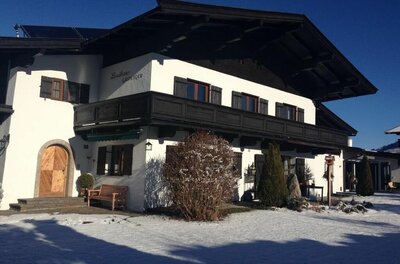 Zweistöckiges weißes Landhaus mit dunklem Holzbalkon und Schneefläche im Vordergrund bei blauem Himmel.