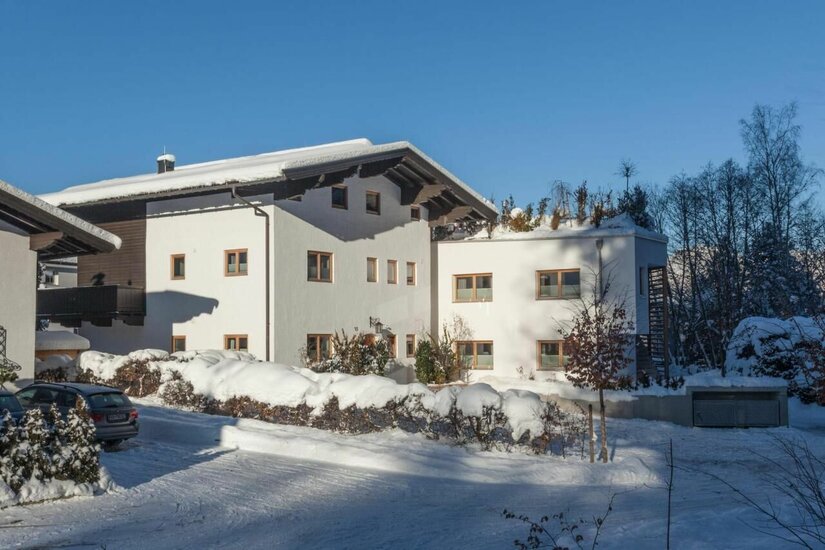 KitzApart Kitz Winteransicht Weißes, mehrstöckiges Alpenhaus mit schneebedecktem Satteldach unter blauem Himmel im Winter.