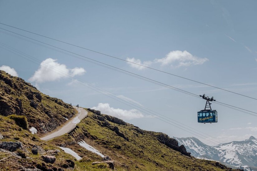 Eine blaue Seilbahnkabine schwebt vor einem Berghang mit Wanderweg und schneebedeckten Gipfeln im Hintergrund.