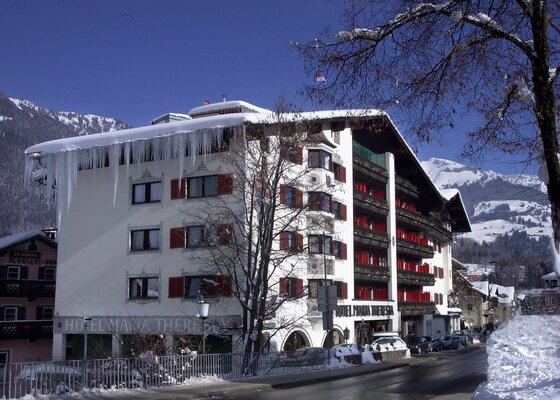 Mehrstöckiges Hotelgebäude mit Schneelast und Eiszapfen am Dachrand vor verschneiten Bergen.
