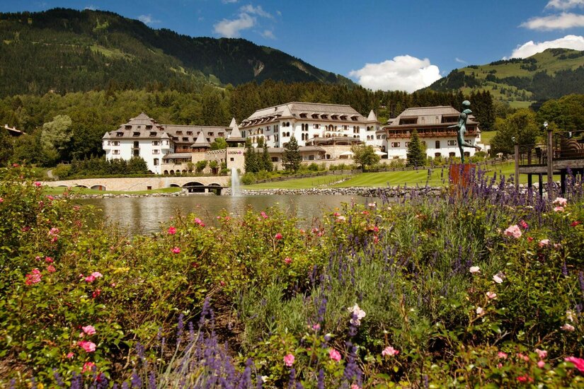 Großes Hotelgebäude hinter einem See mit Blumenbeeten im Vordergrund und bewaldeten Bergen im Hintergrund.