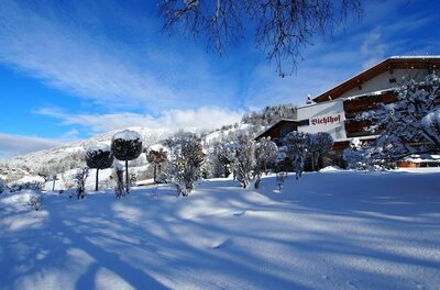 Verschneite Gartenanlage vor dem Hotel Bichlhof mit Bergen im Hintergrund unter blauem Himmel.