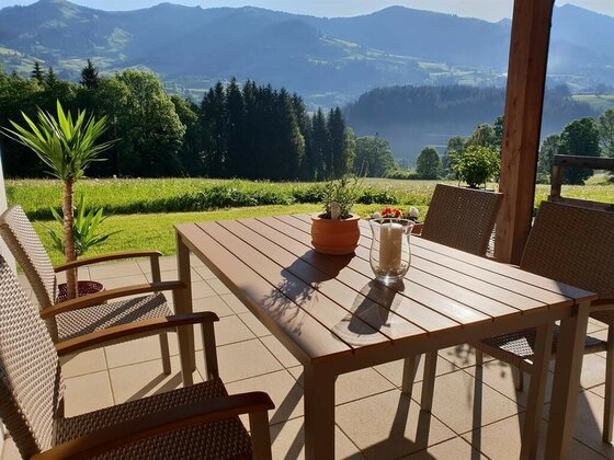 Holztisch und Flechtstühle auf einer Terrasse mit Blick auf bewaldete Berge.