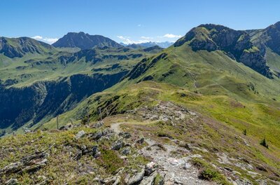 Ein schmaler Wanderpfad führt über einen grasbewachsenen Gebirgskamm mit Blick auf umliegende Berggipfel.