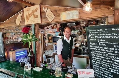Ein Mann in Trachtenweste steht hinter einem hölzernen Tresen mit Getränkekarte und Gläsern.