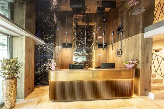 Ein moderner Empfangsbereich mit einem eleganten, goldenen Empfangstresen. Die Wände sind aus Holz und Stein, dekoriert mit Pflanzen und stilvollen Beleuchtungselementen.